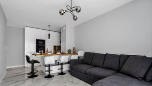 A modern living room with a grey sofa and a dining area with white chairs and a wooden table. - Białołęka Apt - 61m2, Parking & Balcony - by Rentujemy (Warszawa)