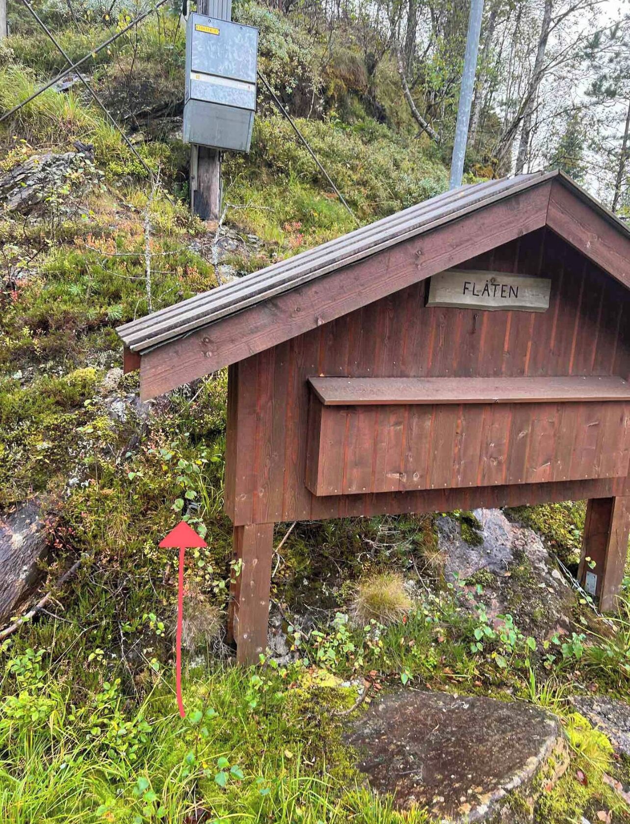 Une structure en bois portant l’inscription « FLÄTEN », avec un toit incliné, située sur une colline herbeuse parsemée de rochers et de végétation.
