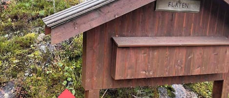 Une structure en bois portant l’inscription « FLÄTEN », avec un toit incliné, située sur une colline herbeuse parsemée de rochers et de végétation.