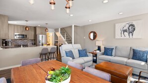 Una cocina moderna con electrodomésticos de acero inoxidable, un comedor con mesa de madera y un frutero, un conjunto de sofá gris y una escalera que lleva a un nivel superior.