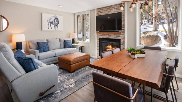 Una sala de estar moderna con un sofá, un reposapiés y una mesa de comedor con sillas. Cuenta con una chimenea, un televisor montado en la pared y un ventanal con vistas al exterior.