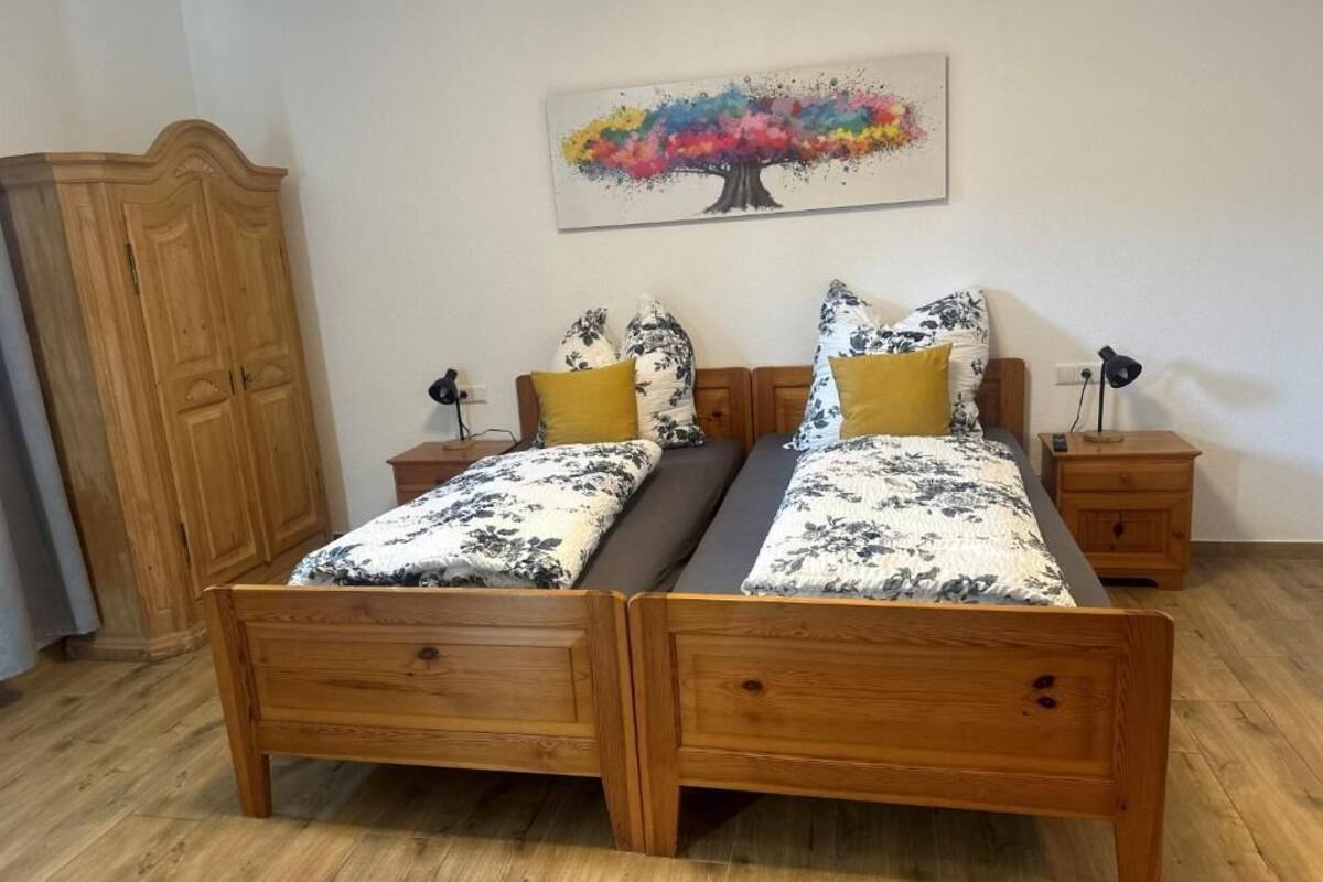 Un lit double avec une literie à motifs floraux, deux tables de chevet, une armoire en bois et un tableau coloré représentant un arbre accroché au mur.