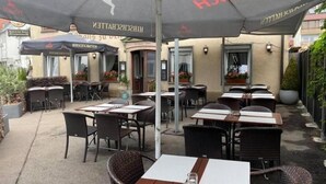 An outdoor café with tables and chairs under umbrellas, a building entrance, and potted plants. - Hotel 7 Winde (Spaichingen)