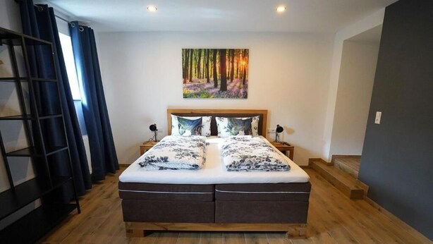 A bedroom with a wooden bed, two nightstands, a painting of a forest, and a wooden floor. - Hotel 7 Winde (Spaichingen)