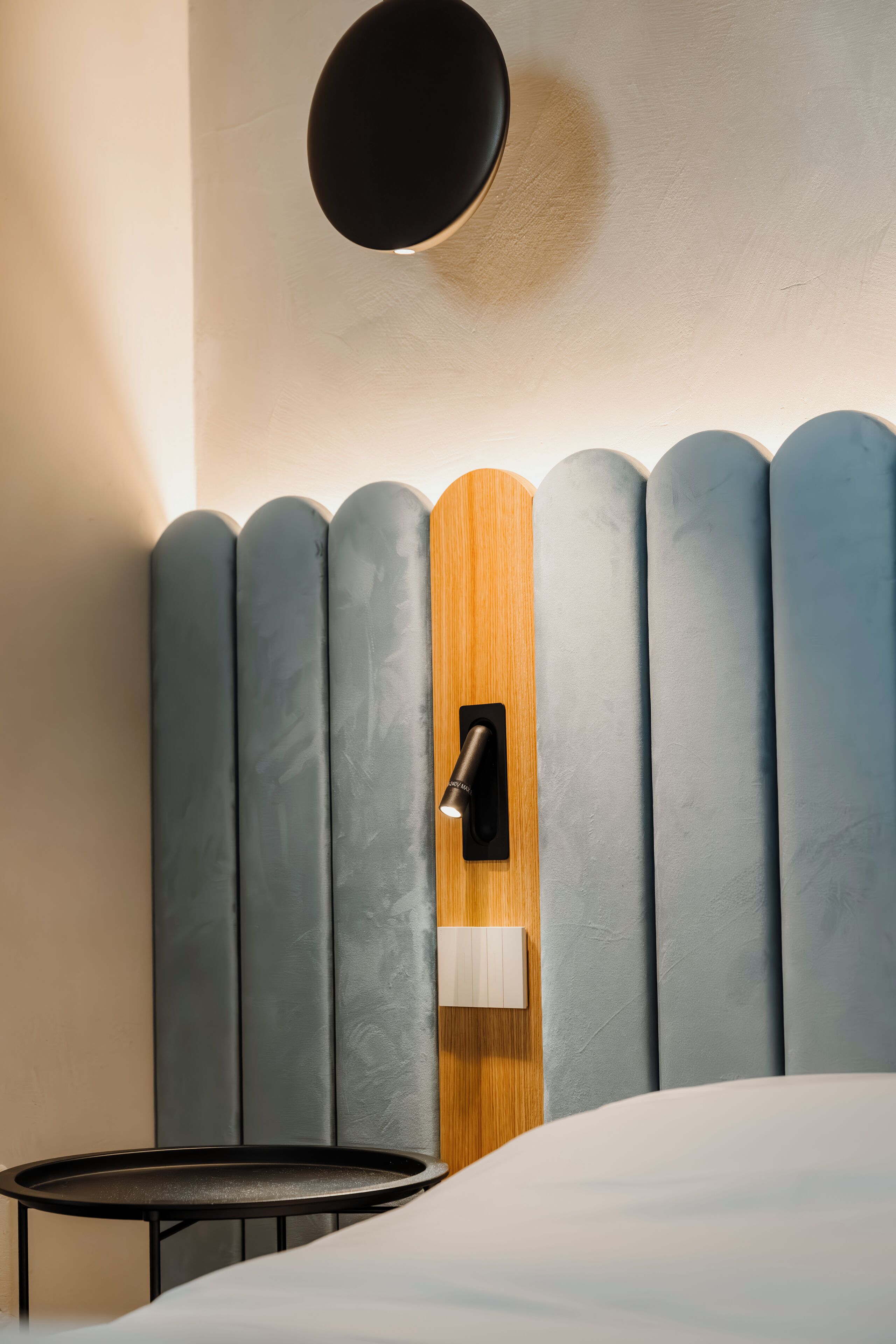 A modern hotel room with a bed, a small round table, and a unique headboard with vertical panels.