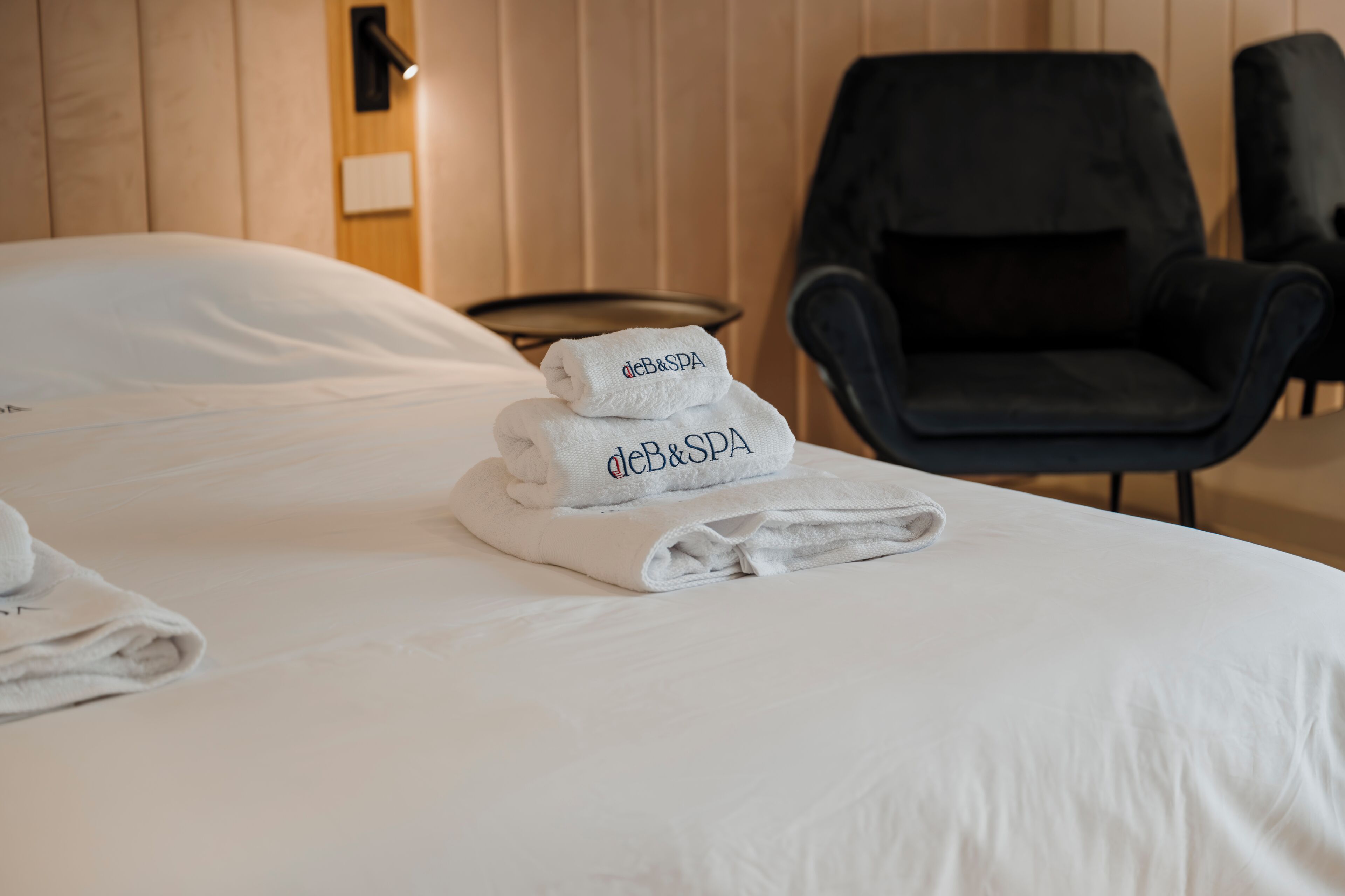 A neatly made bed with folded towels and a black chair in a room with wooden paneling.