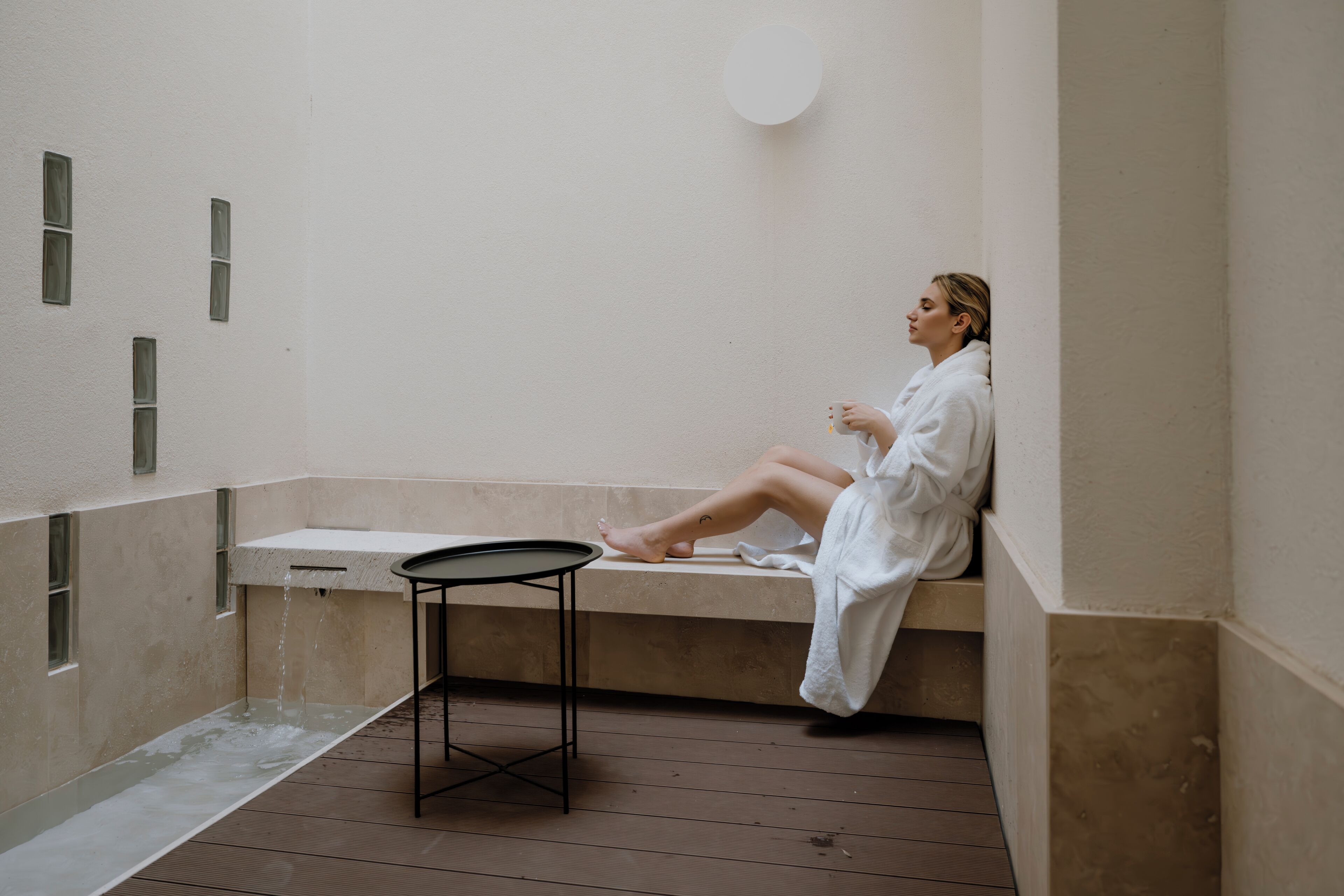 A person in a white bathrobe sitting on a bench in a room with a bathtub, a small table, and a wall-mounted circular light fixture.