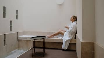 A person in a white bathrobe sitting on a bench in a room with a bathtub, a small table, and a wall-mounted circular light fixture.