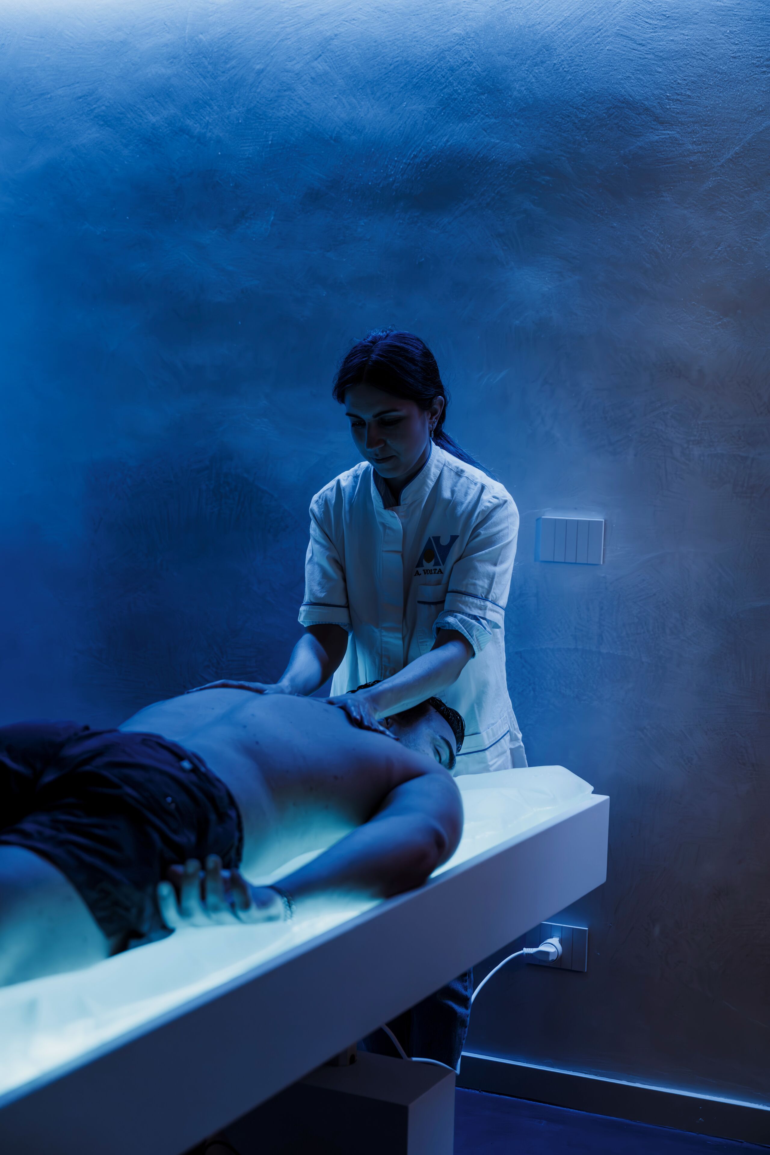 A person lying on a bed in a room with a blue hue, a wall-mounted light switch, and a visible electrical outlet.