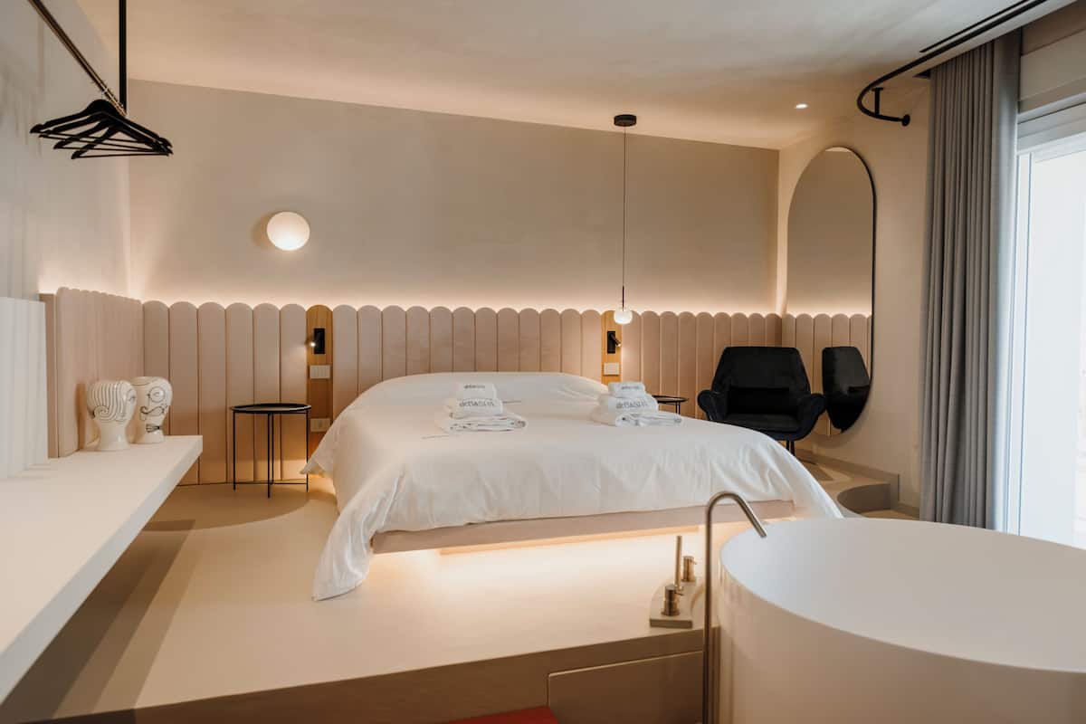 Une chambre d’hôtel moderne avec un grand lit, un miroir rond, une chaise noire, une petite table et une baignoire.