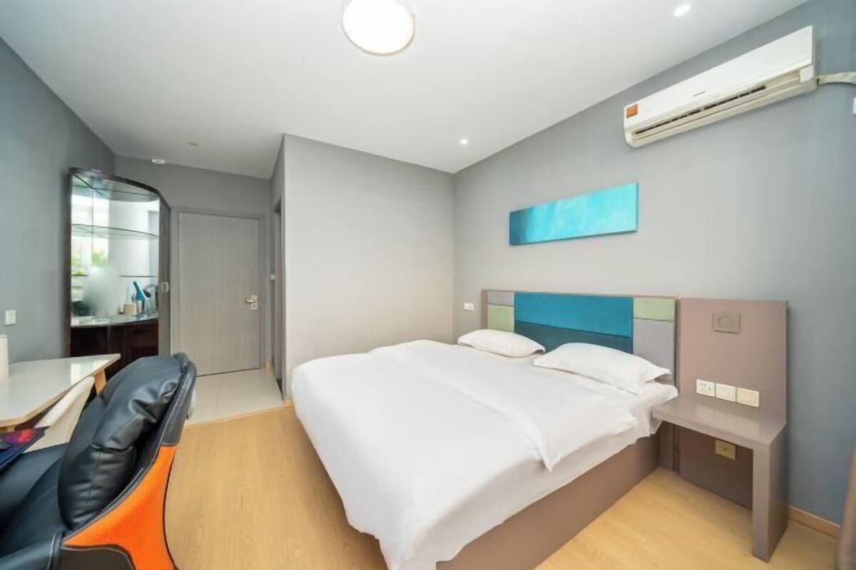 A hotel room with a bed, a desk, a chair, a bedside table, and an air conditioning unit.