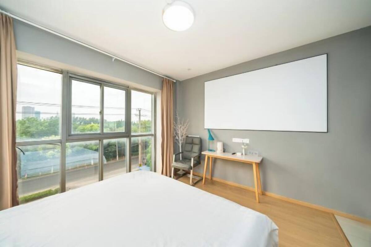 A modern hotel room with a large bed, a desk, and a view of the outdoors through large windows.