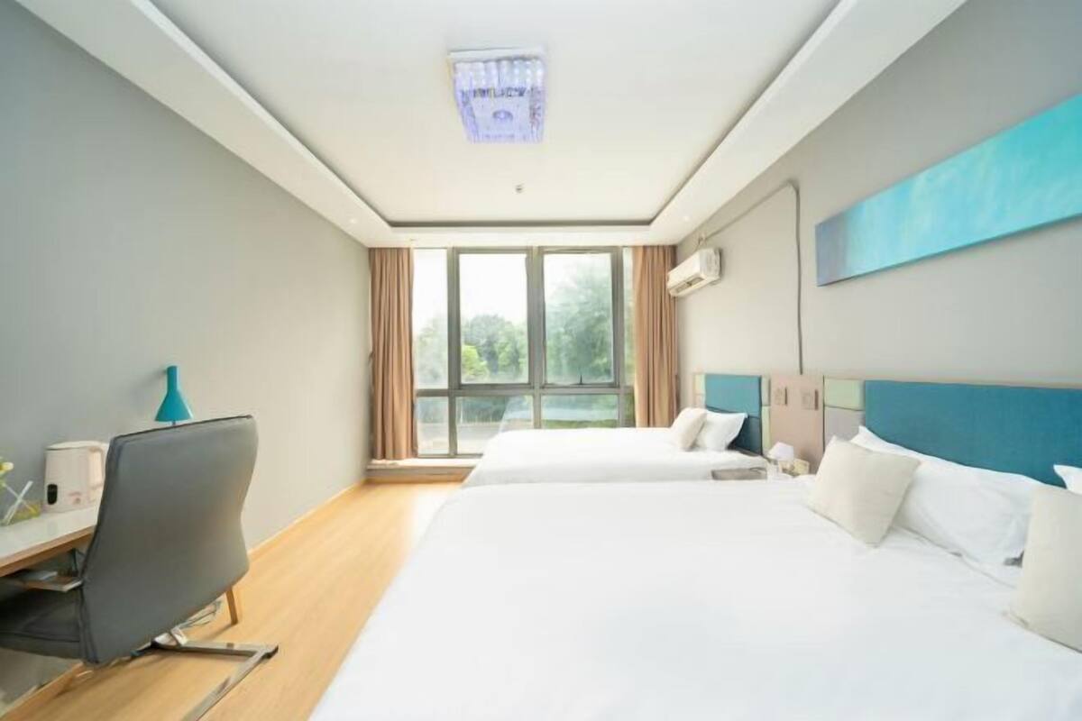 A hotel room with two beds, a desk with a chair, a window with curtains, and a ceiling light fixture.