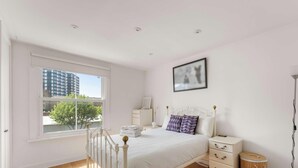 A bedroom with a large window, a bed, a bedside table, a nightstand, and a framed picture on the wall.