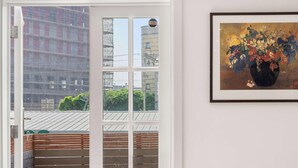A room with a door leading to an outdoor area, a framed painting of a floral arrangement on the wall, and a view of buildings through the door.