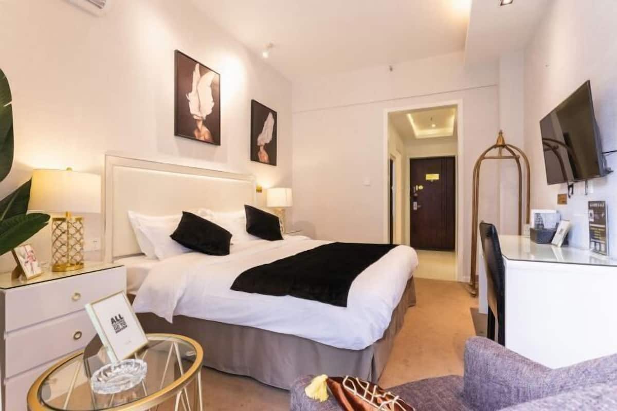 A modern hotel room with a neatly made bed, a glass coffee table, a comfortable armchair, and a flat-screen TV mounted on the wall.