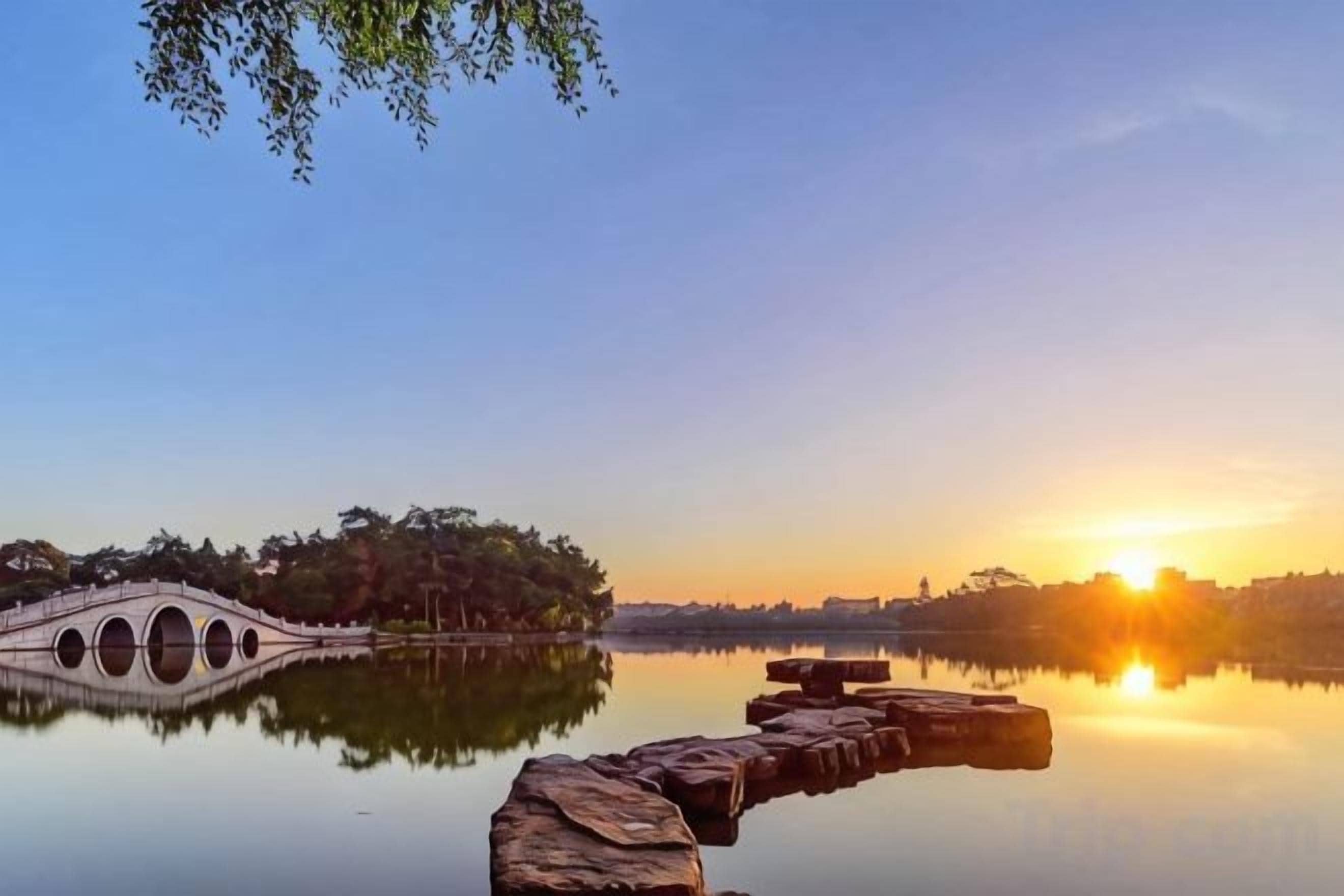 A calm lake with a stone bridge and a sunset in the background.