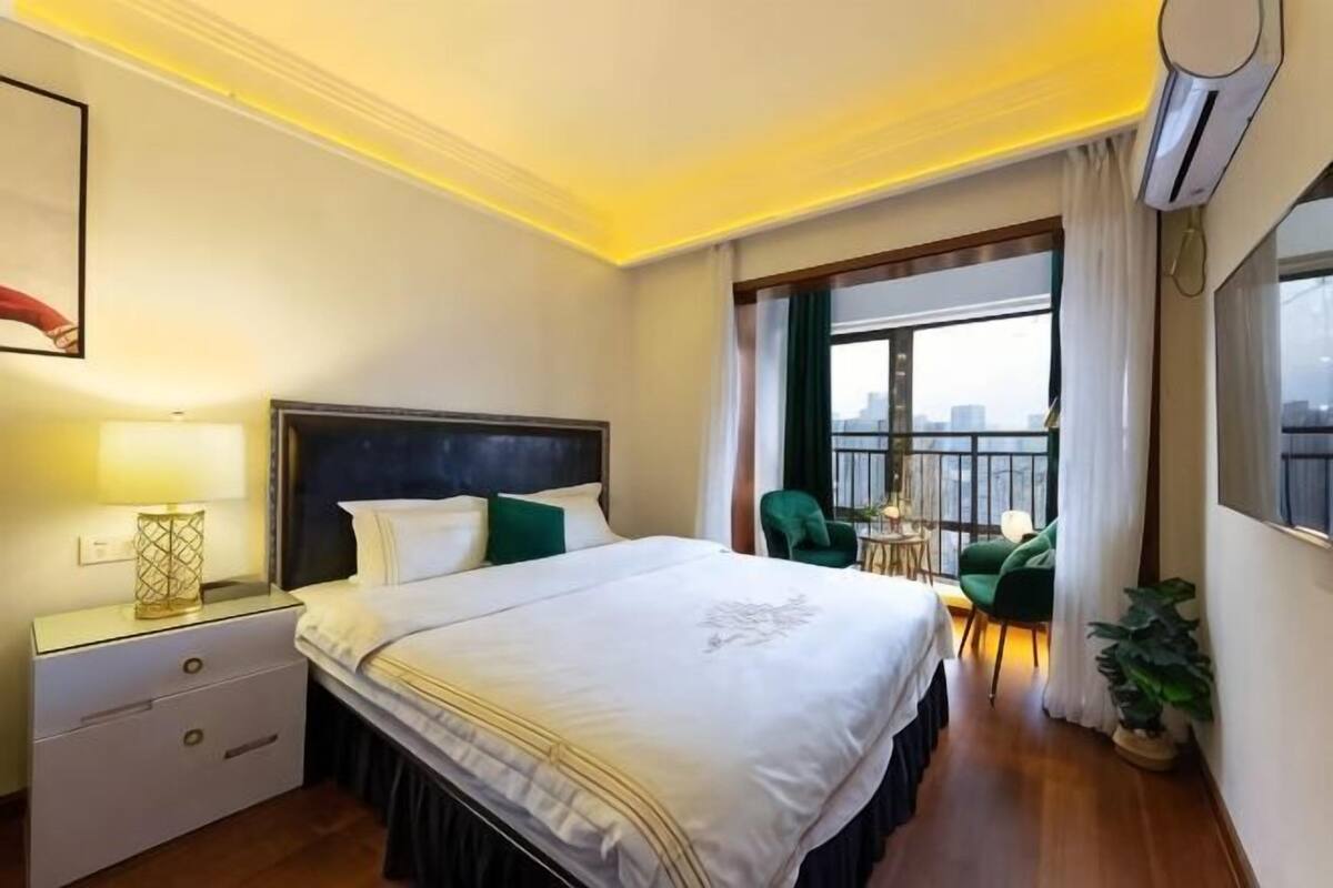 A hotel room with a bed, bedside table, lamp, chair, potted plant, and a view of the city through a window.