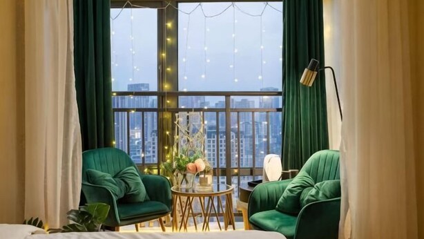 Een kamer met een groot raam, twee groene stoelen, een kleine tafel met een vaas en een lamp.