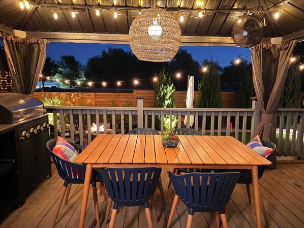 Un patio al aire libre con una mesa y sillas de madera, una parrilla y luces colgantes.