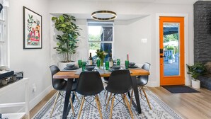 A dining area with a wooden table set for four, surrounded by black chairs, a potted plant, and a framed picture on the wall.