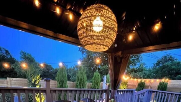 A wooden deck with a table and chairs, a hanging light fixture, and a view of trees and a fence.