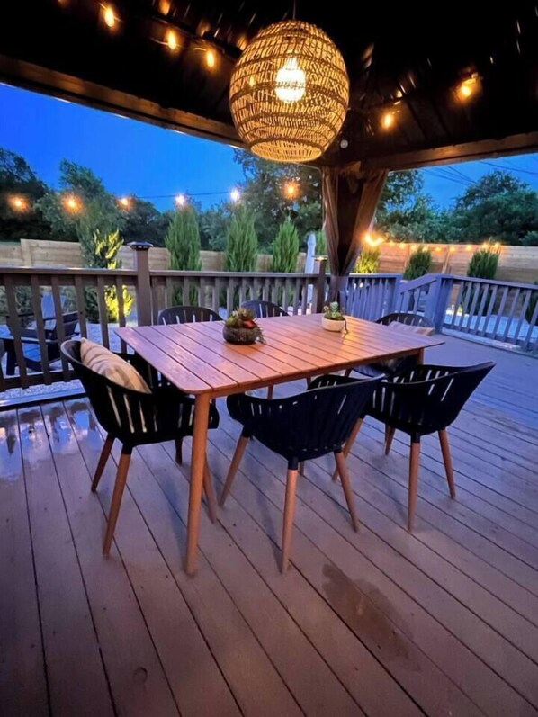 A wooden deck with a table and chairs, a hanging light fixture, and a view of trees and a fence.