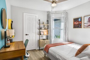 Un dormitorio con cama, escritorio, silla, estante con objetos, ventana con cortinas y dos obras de arte en la pared.