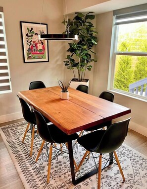 A dining area with a wooden table, black chairs, a hanging light fixture, a potted plant, and a framed picture on the wall.
