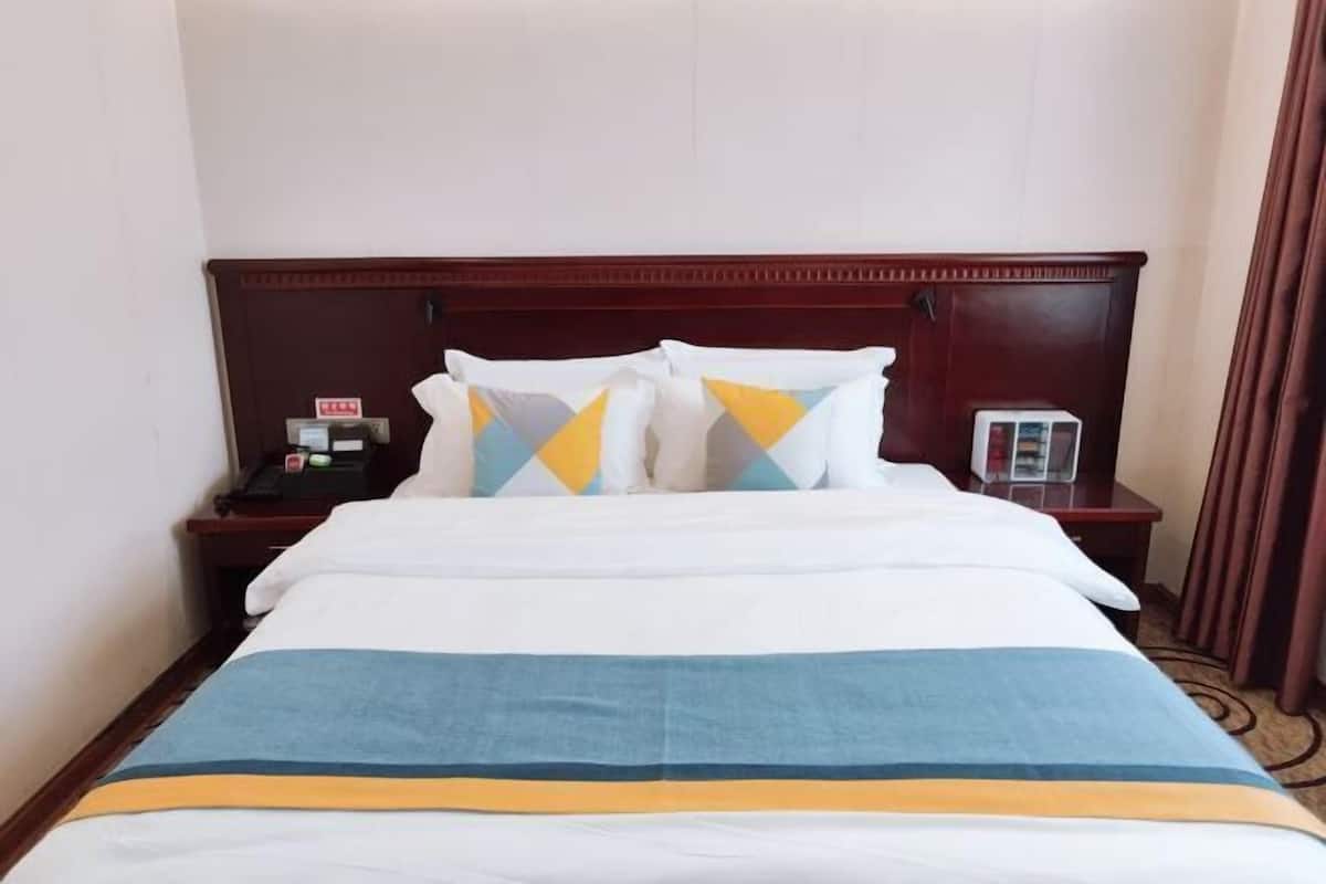 Una habitación de hotel con cabecera de madera, cama con ropa de cama blanca y almohadas de colores, mesita de noche con un teléfono y un vaso, y cortina marrón.