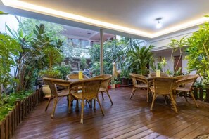 Un patio interior con mobiliario de mimbre, rodeado de abundante vegetación y vistas a un edificio.