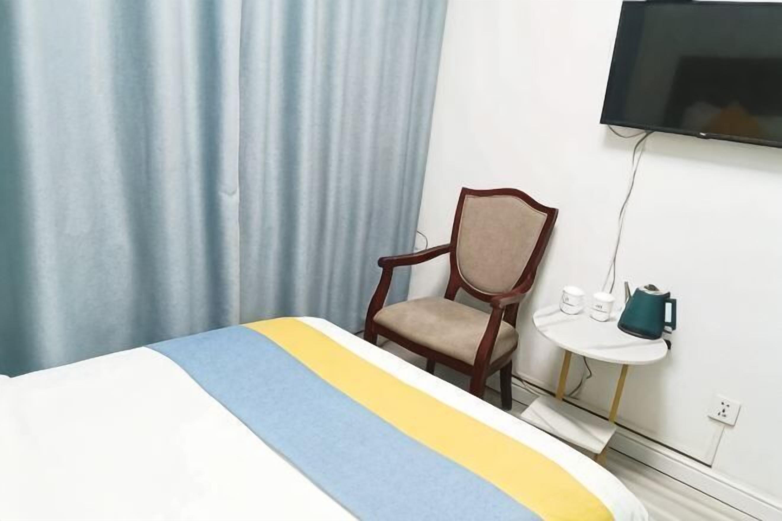 A hotel room with a bed, a chair, a small table with a kettle, and a TV mounted on the wall.