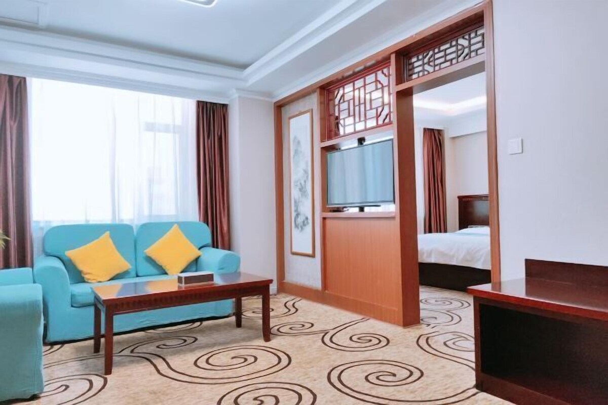A hotel room with a blue sofa, a wooden coffee table, a bed with white bedding, and a television on a wooden stand.