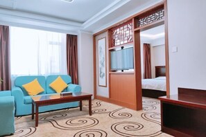 A hotel room with a blue sofa, a wooden coffee table, a bed with white bedding, and a television on a wooden stand.