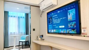 A hotel room with a large TV mounted on the wall, a small table with a chair, and a window with curtains. - fengyuhotel (Shanghai)