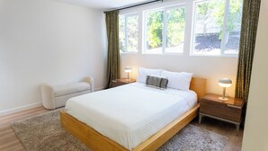 Une chambre moderne avec un grand lit, deux tables de chevet avec des lampes, un banc et de grandes fenêtres avec des rideaux.