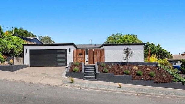 Une maison moderne à deux étages, dotée d’un garage, d’une entrée avec quelques marches et d’un jardin aménagé avec des arbustes et un petit arbre.