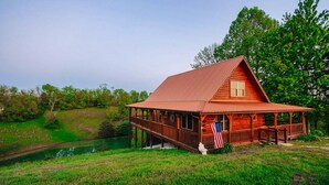 Une cabane en bois dotée d’un porche, entourée de verdure et bordée d’un petit étang.