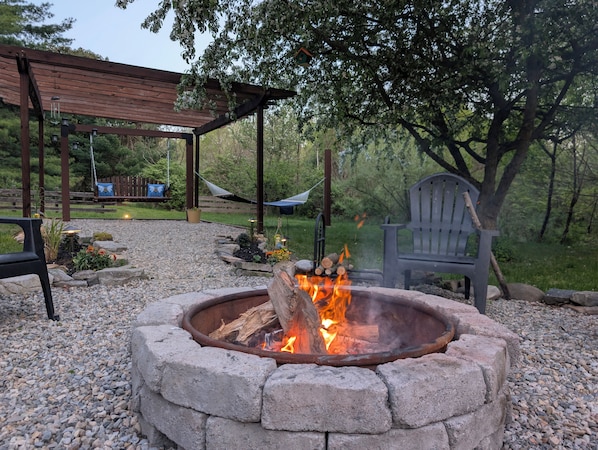 A fire pit surrounded by stone, a hammock, a wooden pergola, and a chair in a garden setting.