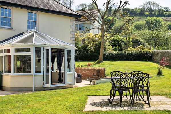 A conservatory with glass doors, a patio with chairs, and a well-maintained lawn. - Cottage in Millbrook Near Rame Peninsula (Torpoint)