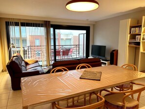 A living room with a sofa, a dining table with chairs, a TV, and a bookshelf. - Cozy 2-bedroom Apartment Near Koksijde Beach (Koksijde)