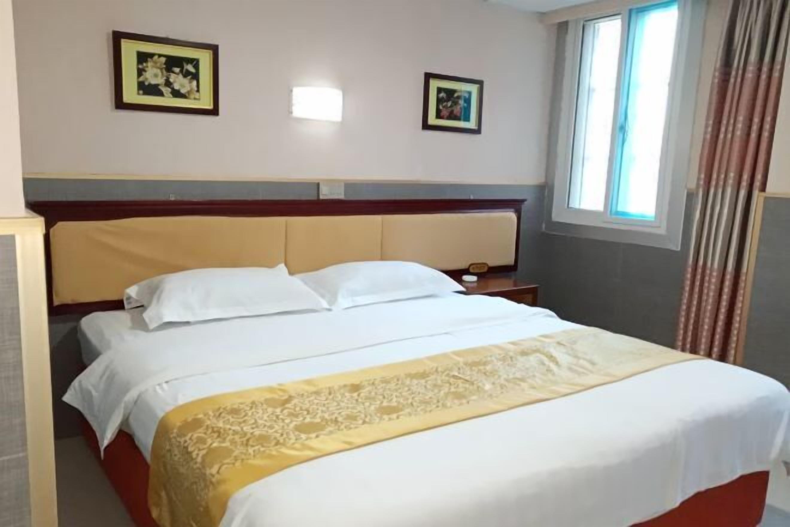 A hotel room with a bed, two framed pictures on the wall, a window with curtains, and a bedside table.