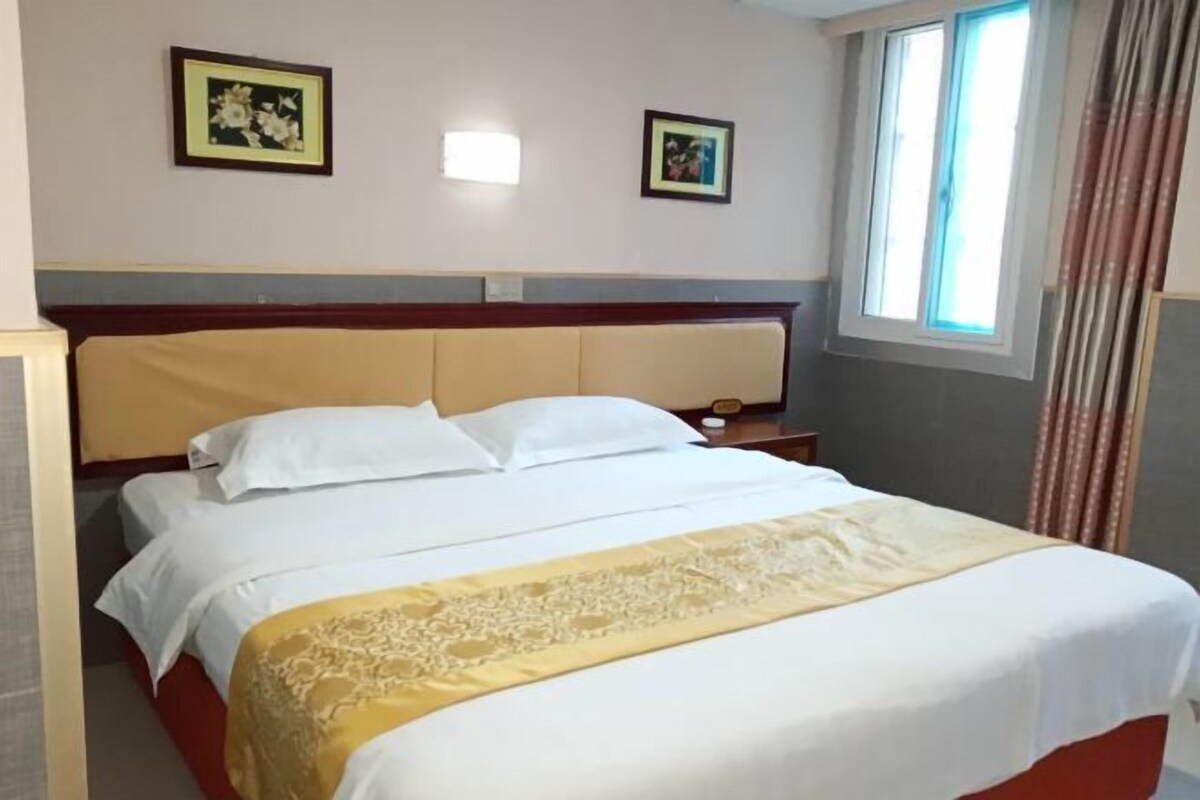 A hotel room with a bed, two framed pictures on the wall, a window with curtains, and a bedside table.