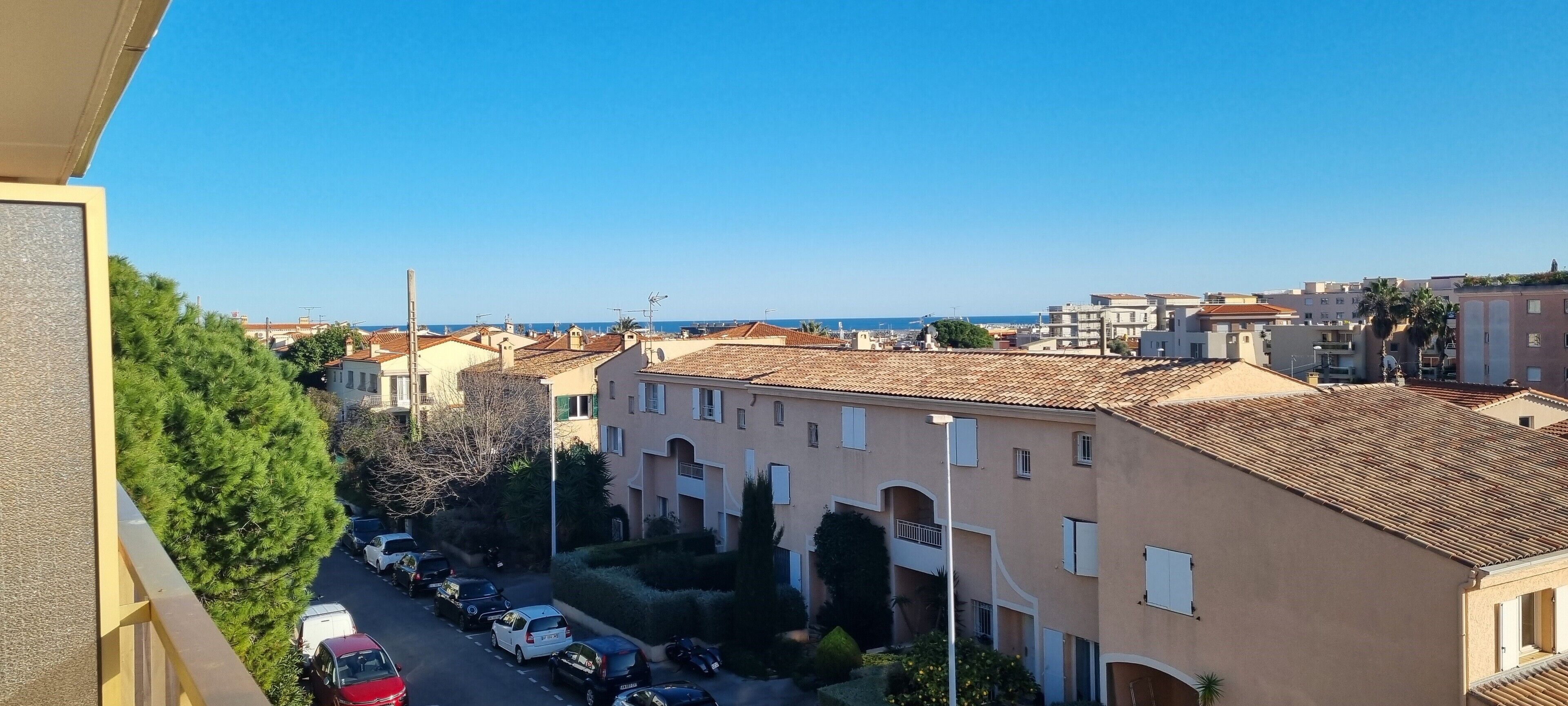 A residential area with multi-story buildings, a clear sky, and a view of the sea in the distance.
