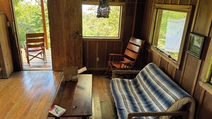 Interior de una cabaña de madera con una mesa, un banco y una silla. Se ve una puerta que da al exterior y una ventana con vistas a la vegetación.