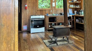 Interior rústico de una cabaña de madera con una zona de cocina que incluye estufa, horno y chimenea de leña. Hay dos ventanas con vistas al follaje exterior, y un extintor de incendios montado en la pared.