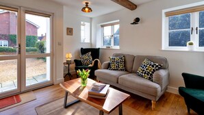 Een woonkamer met een bank, salontafel en stoelen, grote ramen met zonwering, en een houten balk.