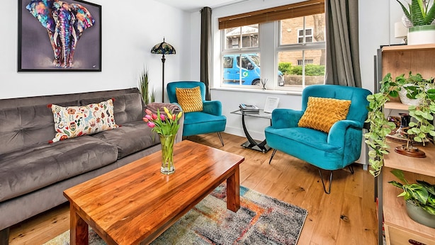 A modern living room with a grey sofa, a wooden coffee table, two blue armchairs, a colorful elephant painting, and a view of a blue car outside. - Apartment in Harrogate Near Yorkshire Dales (Harrogate)