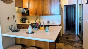A kitchen with wooden cabinets, a white countertop, and a small island with stools. There is a microwave, toaster, and various kitchen utensils on the counter.