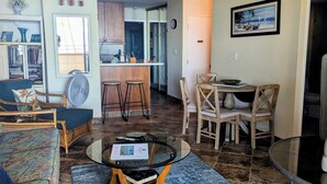 A living room with a sofa, a coffee table, and a dining area with chairs and a table. There is a kitchen in the background with cabinets and a window.